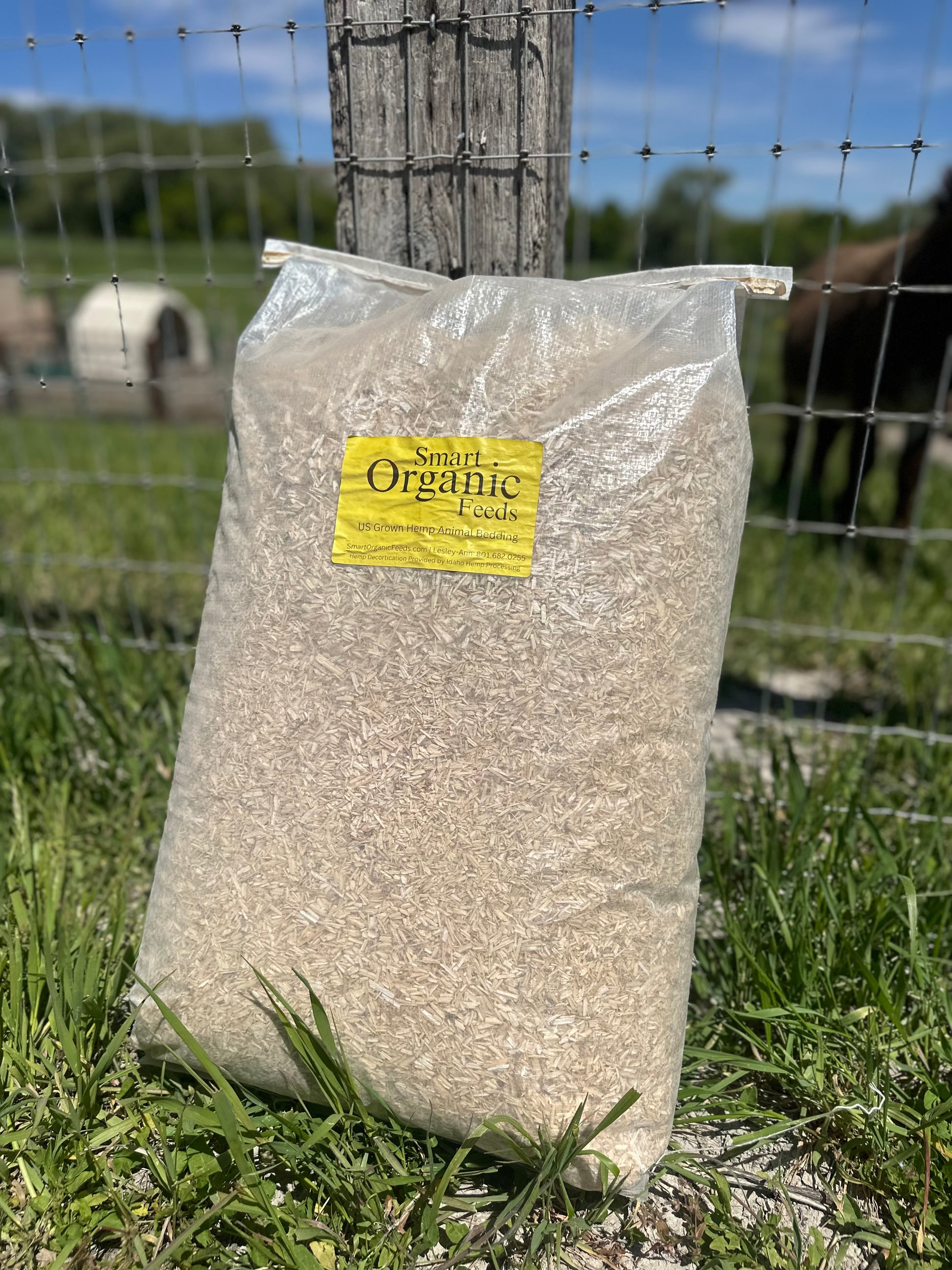 A large clear plastic bag filled with hemp bedding sitting on grass in front of a wire fence and wooden post outdoors. 
