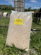 A large clear plastic bag filled with hemp bedding sitting on grass in front of a wire fence and wooden post outdoors. 